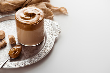 delicious Dalgona coffee in glass near cloth, brown sugar and spoon on silver platter on white background