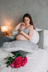 Wall Mural - pregnant girl model woman sitting on bed in tracksuit near pink peonies with hug pillow