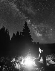 Wall Mural - side view of female tourist having a rest by campfire on background of spruce forest under starry sk