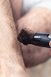 Dłoń mężczyzny trzyma maszynkę elektryczną do strzyżenia włosów i obcina długie ciemne włosy na swoich nogach.