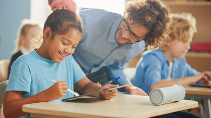 Wall Mural - Elementary School Computer Science Class: Smart Girl Uses Digital Tablet Computer, Friendly Teacher Helps Her by Explaining Lesson and Asignment. Children Getting Modern Education