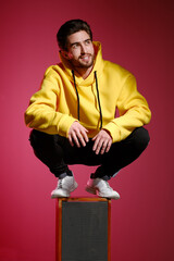 Wall Mural - A young man of 25-30 years old in a yellow sweatshirt emotionally standing on an old music column on pink wall background. 