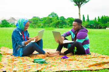 young black couples sitting down in a park, using their laptops to surf the internet