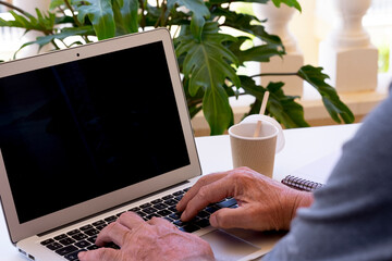 Senior man using laptop, typing on keyboard searching web, browsing information, working from home terrace  on a white table