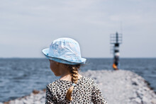 A Little Girl With Her Back In A Blue Panama Hat Looks Into The Distance To The Sea On A Rocky Shore Near The Lighthouse.