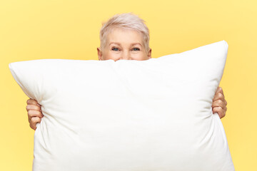 Bedding, sleep and bedtime concept. Cropped shot of beautiful middle aged female with short hair hiding herself behind white large feather pillow with copy space for your text or advertisement