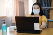 © EduLife Photos - A teenager student wears surgical mask and studies online via laptop during COVID-19 pandemic. University student girl watches online classes and writing a syllabus in notebook. Stock photo.