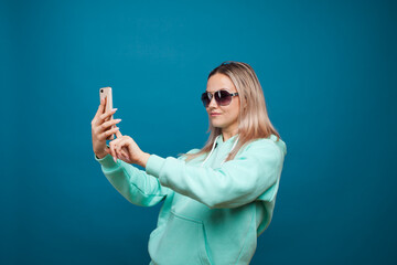 Wall Mural - Cheerful blonde with a mobile phone. Portrait of a young positive woman in a blue hoodie