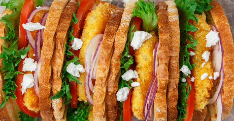 Wall Mural - Gourmet sandwiches with fried fish (Pollock, cod, hake, nototenia, perch), lettuce, feta cheese, tomatoes and red onion close-up. Tasty snack. Picnic. Culinary background. Selective focus, banner