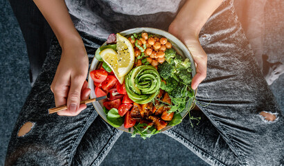 Wall Mural - Woman in jeans holding Buddha bowl with salad, baked sweet potatoes, chickpeas, broccoli, greens, avocado, sprouts in hands. Healthy vegan food, clean eating, dieting, top view