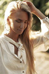 Wall Mural - Charming tanned attractive woman in white cotton blouse poses with closed eyes outside. Blonde girl touches hair and walks outdoors.