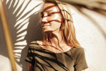 Wall Mural - Pretty blonde long-haired girl in dark stylish top and with gold earrings smiles with closed eyes and poses near white wall with palm leaf shadow.