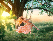 Beautiful Happy Woman Nymph Sitting On Swings. Magical Fantasy Swing. Princess Long Peach Color Orange Silk Vintage Dress Fluttering Wind. Blond Hair Fly In Motion. Tree Sunshine Green Grass Forest