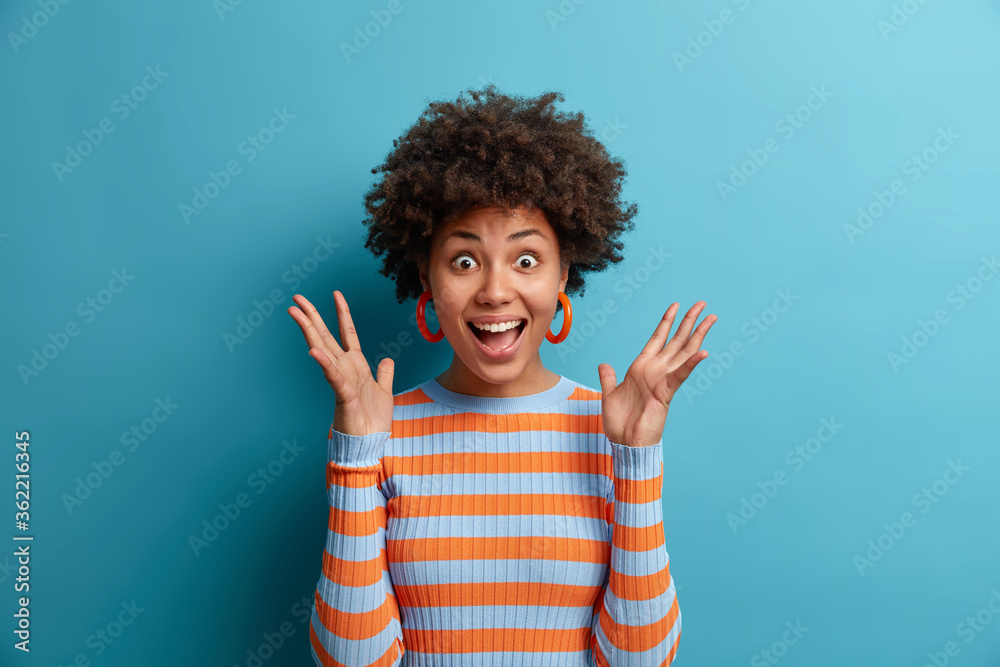 Portrait of delighted cheerful ethnic woman reacts on awesome gift, raises palms and stares with eyes full of happiness, cannot believe in her great success, dressed in casual wear, poses indoor - obrazy, fototapety, plakaty 