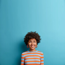 Vertical Shot Of Good Looking Afro American Woman Looks Above With Broad Smile, Notices Nice Offer Or Sale, Feels Happy Dressed In Casual Striped Jumper Isolated On Blue Background. Copy Space Upwards