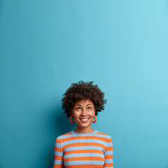 Wall Mural - Vertical shot of good looking Afro American woman looks above with broad smile, notices nice offer or sale, feels happy dressed in casual striped jumper isolated on blue background. Copy space upwards