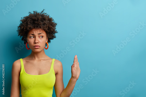 Serious dissatisfied woman with curly hair makes stop gesture, pulls palm forward away, asks not to come, shows warning sign, tells I refuse, shows definitive no, warns and declines conflict