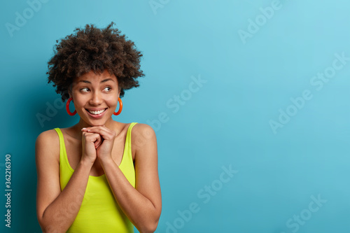Horizontal studio shot of cheerful African American woman keeps hands under chin and looks pleasantly aside, being in good mood, chuckles as gets compliment from boyfriend, empty space on blue wall