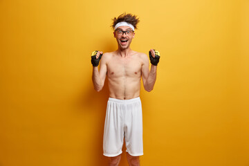 Funny positive skinny man raises hands with clenched fists, feels very happy and motivated, wears white headband, sport gloves and shorts, poses against vivid yellow background, ready to train muscles