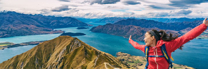 Hiker jumping of joy funny - panoramic banner of woman hiking in New Zealand laughing having fun, joyful and aspirational and carefree at Roys Peak, South Island, New Zealand.