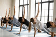 © fizkes - Group of young fit mixed race people standing in row in side plank with raised arm on floor mat, strengthening core muscles in Vasisthasana position, improving stability, working out in yoga studio.