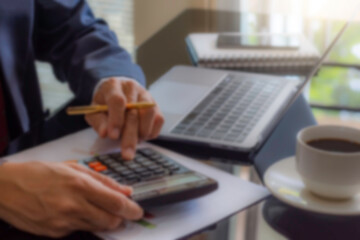 Wall Mural - Blurred image of businessman in suit hand using calculator and work on laptop computer with data chart paper, notebook, phone and cup of coffee on the desk at modern office. Tax time concept.