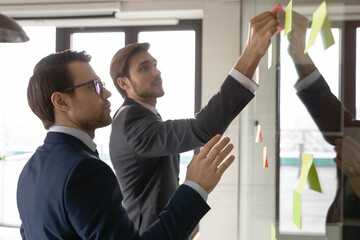 Wall Mural - Motivated male colleagues write ideas on sticky post in notes attach to glass wall in office, focused Caucasian man employees develop business plan, engaged in creative thinking in boardroom