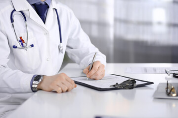 Unknown male doctor sitting and working with clipboard of medication history record in clinic at his working place, close-up. Young physician at work. Perfect medical service, medicine concept