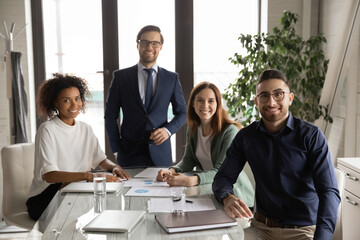 Portrait of smiling multiracial young businesspeople sit at desk in office look at camera poising together, happy multiethnic colleagues show unity and motivation at workplace, teamwork concept