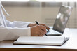 © fizkes - Crop close up of male doctor in white uniform sit at desk work on laptop fill patient anamnesis in medical journal, man GP or physician make notes in paper register, use computer, healthcare concept