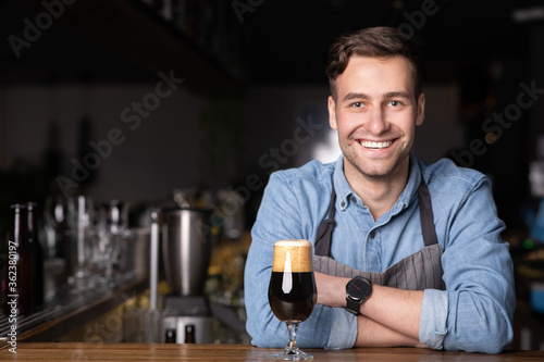 Modern smiling barman in apron with crossed arms in interior of bar