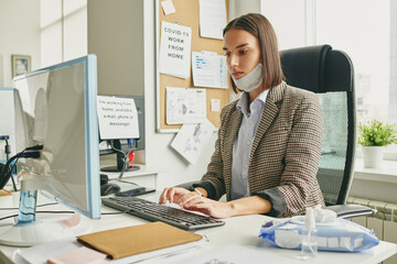 Poster - Young serious businesswoman with protective mask on chin typing in office