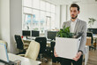 © pressmaster - Sad young man carrying box with green plant and documents while leaving office