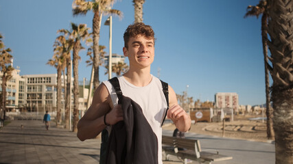 Handsome fit guy with backpack happily walking along seaside after workout