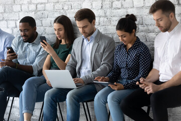 Wall Mural - Group of multi racial young people focused in gadgets, applicants sit in row await in line for job interview use devices spend time in internet ignoring of each other, phubbing and employment concept