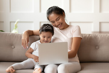 Little cute asian baby sitting on sofa with smiling affectionate vietnamese ethnicity mum nanny, looking at computer screen, watching funny childish cartoons or using early education applications.