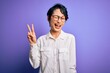 Young beautiful asian girl wearing casual shirt and glasses standing over purple background smiling with happy face winking at the camera doing victory sign with fingers. Number two.