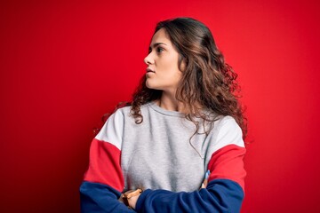 Sticker - Young beautiful woman with curly hair wearing casual sweatshirt over isolated red background looking to the side with arms crossed convinced and confident