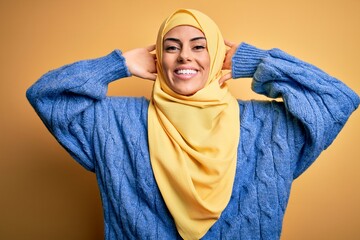 Sticker - Young beautiful brunette muslim woman wearing arab hijab over isolated yellow background relaxing and stretching, arms and hands behind head and neck smiling happy