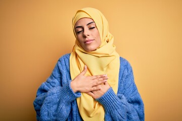 Poster - Young beautiful brunette muslim woman wearing arab hijab over isolated yellow background smiling with hands on chest with closed eyes and grateful gesture on face. Health concept.