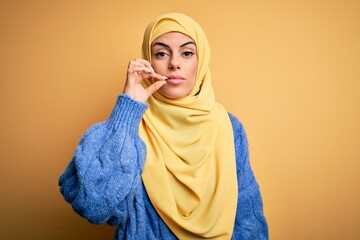 Sticker - Young beautiful brunette muslim woman wearing arab hijab over isolated yellow background mouth and lips shut as zip with fingers. Secret and silent, taboo talking