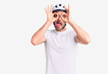 Wall Mural - Young handsome man wearing bike helmet doing ok gesture like binoculars sticking tongue out, eyes looking through fingers. crazy expression.