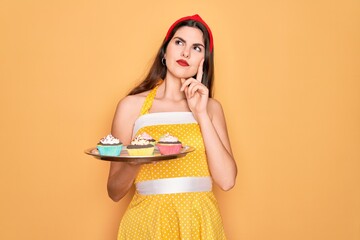 Sticker - Young beautiful pin up woman wearing 50s fashion vintage dress holding tray with cupcakes serious face thinking about question, very confused idea