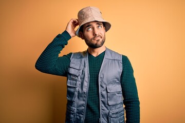 Canvas Print - Handsome tourist man with beard on vacation wearing explorer hat over yellow background confuse and wondering about question. Uncertain with doubt, thinking with hand on head. Pensive concept.