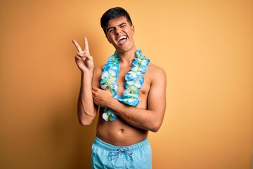 Poster - Young handsome tourist man on vacation wearing swimwear and hawaiian lei flowers smiling with happy face winking at the camera doing victory sign. Number two.