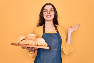 Wall Mural - Young beautiful baker woman with blue eyes wearing apron holding tray with bread very happy and excited, winner expression celebrating victory screaming with big smile and raised hands