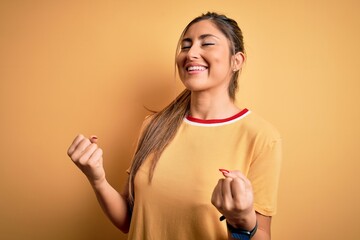 Wall Mural - Young beautiful athletic woman wearing casual t-shirt and ponytail over yellow background very happy and excited doing winner gesture with arms raised, smiling and screaming for success. Celebration.