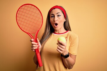 Wall Mural - Beautiful brunette sportswoman playing tennis using racket and ball over yellow background scared in shock with a surprise face, afraid and excited with fear expression