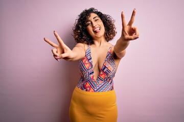 Wall Mural - Young beautiful arab woman on vacation wearing swimsuit and sunglasses over pink background smiling with tongue out showing fingers of both hands doing victory sign. Number two.