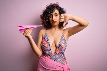 Wall Mural - Young beautiful curly arab woman on vacation wearing swimsuit holding pink paper airplane with angry face, negative sign showing dislike with thumbs down, rejection concept